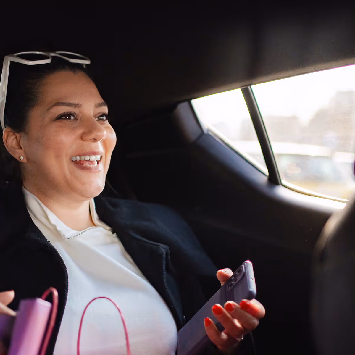 Smiling woman holding a smartphone and a pink bag inside a car near the window.