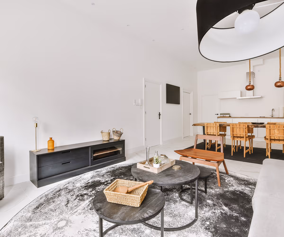 Modern living room with black and gray rug, nested round tables, wooden bench, dining table, and kitchen area.