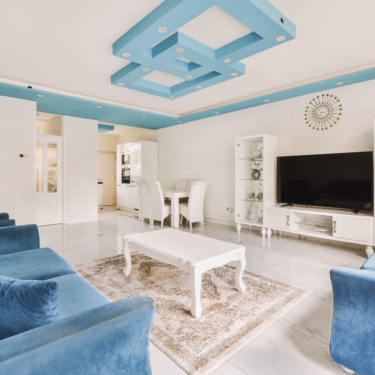 Modern living room with blue sofas, white coffee table, dining set, TV, and decorative ceiling design.