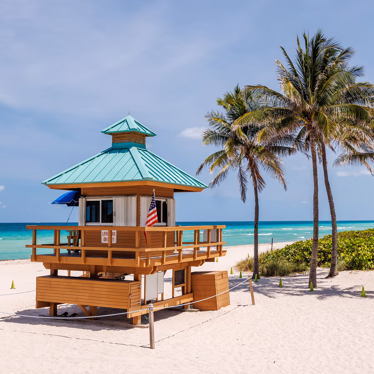 Wooden lifeguard tower with teal roof on sandy beach near palm trees and ocean under blue sky.