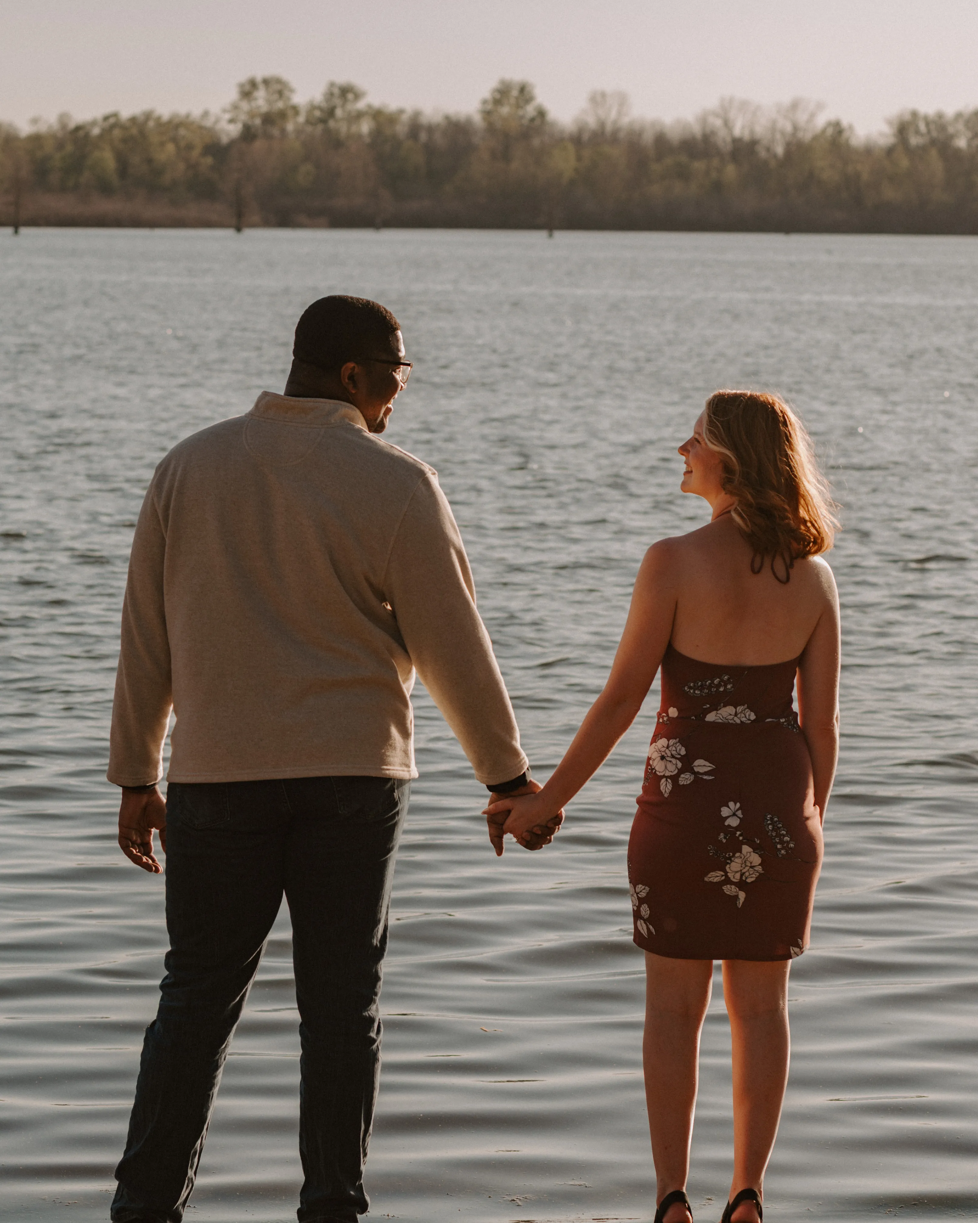 Couple holding hands beside a calm lake with trees on the far shore at sunset.
