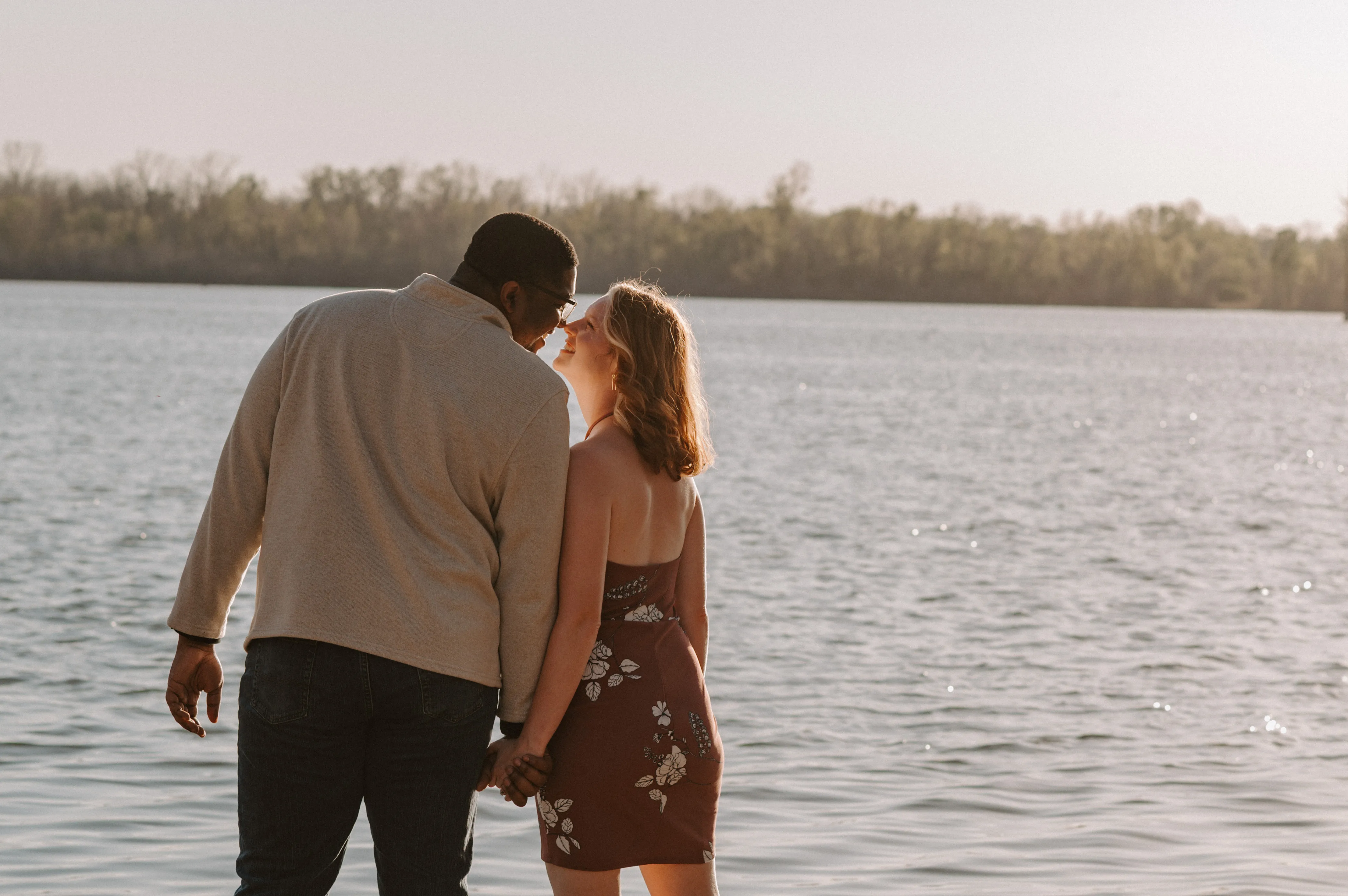 Couple holding hands and smiling by a lake with trees in the background at sunset.