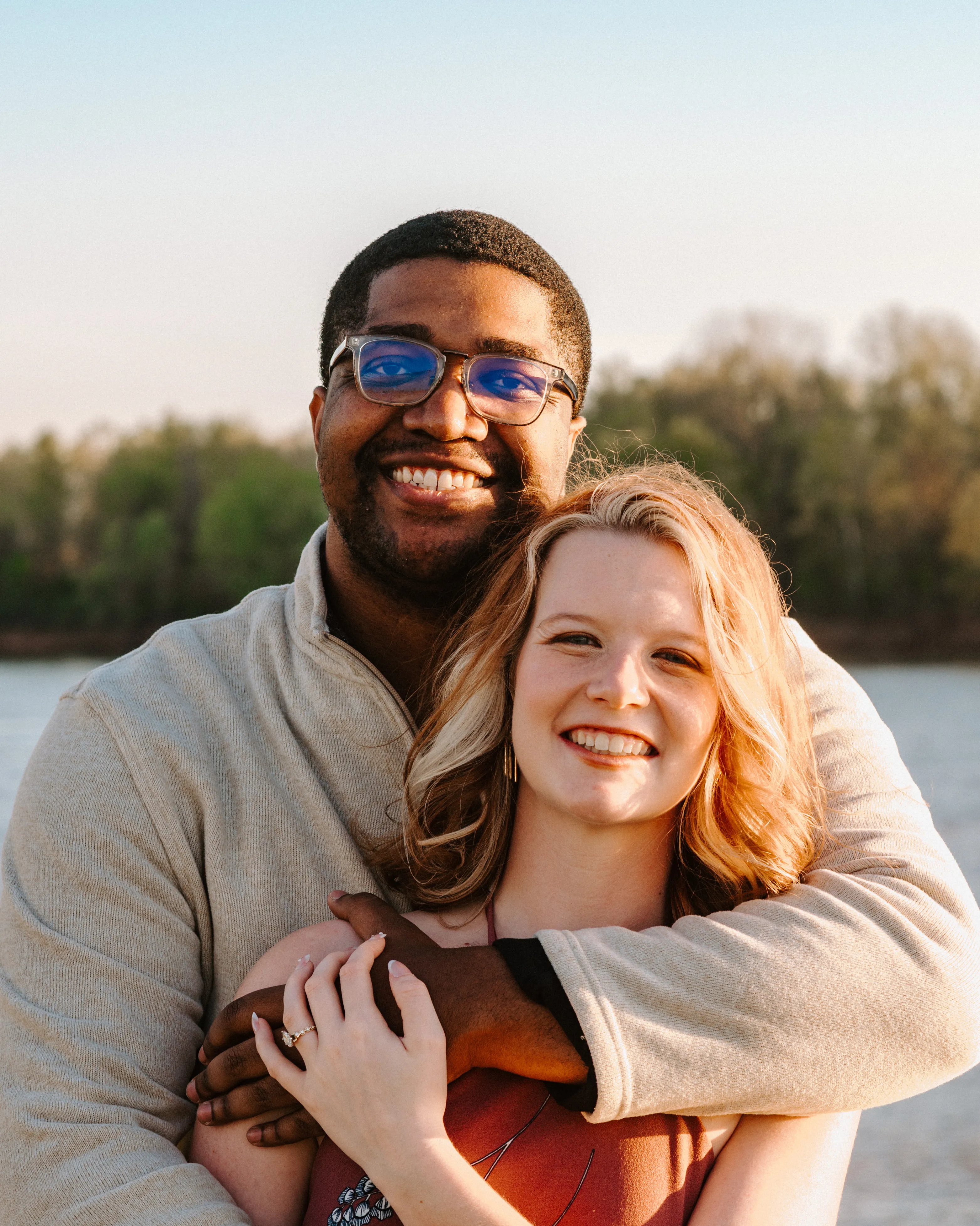 Smiling couple outdoors by water, man hugging woman from behind during golden hour.