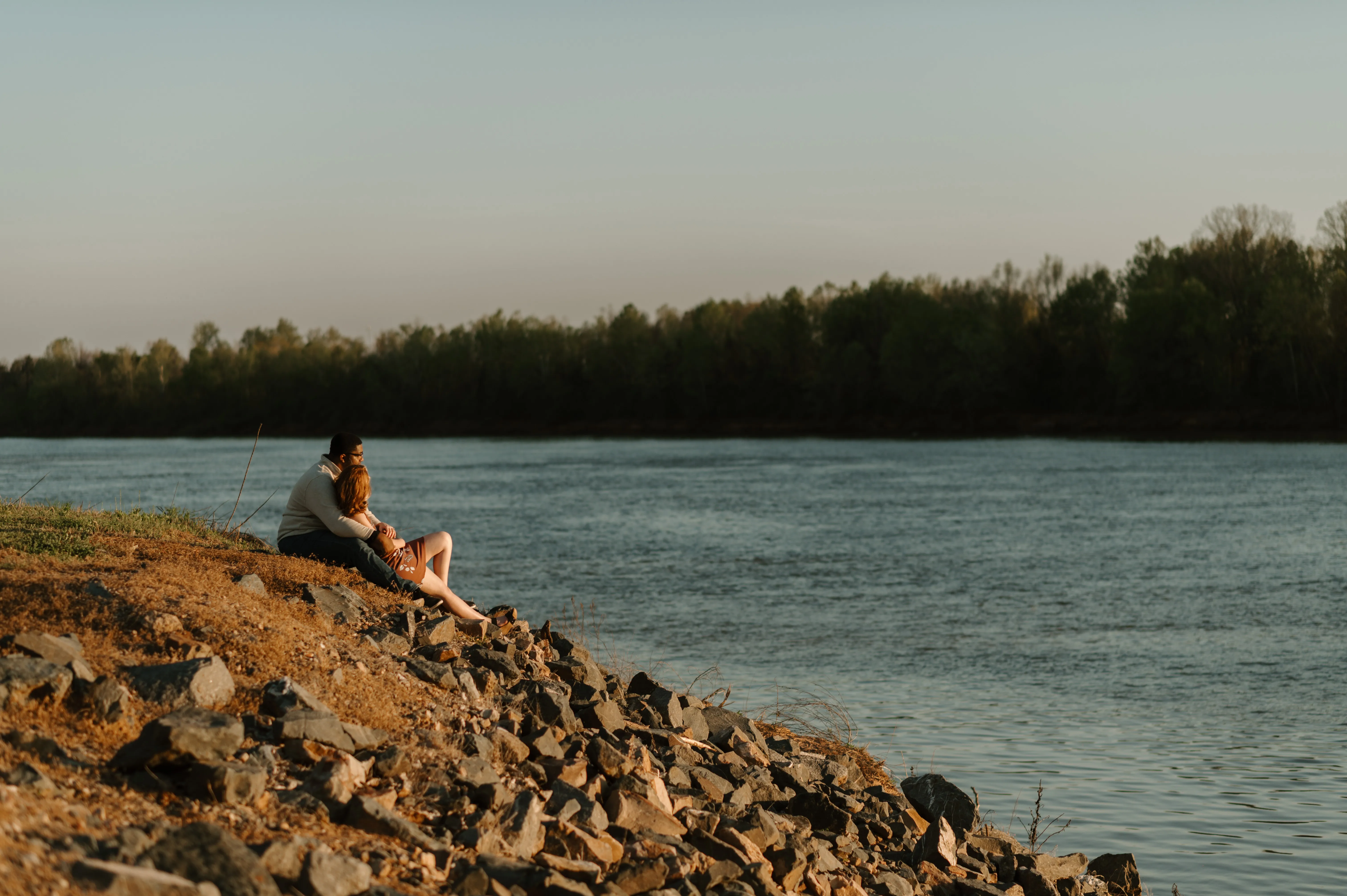 Couple sitting on rocky shore by river, man hugging woman, trees in background under clear sky.