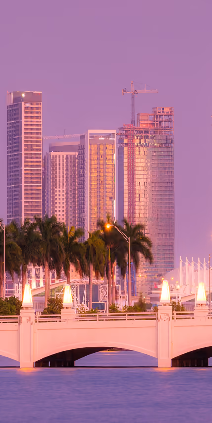 Miami skyline at sunrise with palm trees, high-rise buildings, and a white bridge over water.