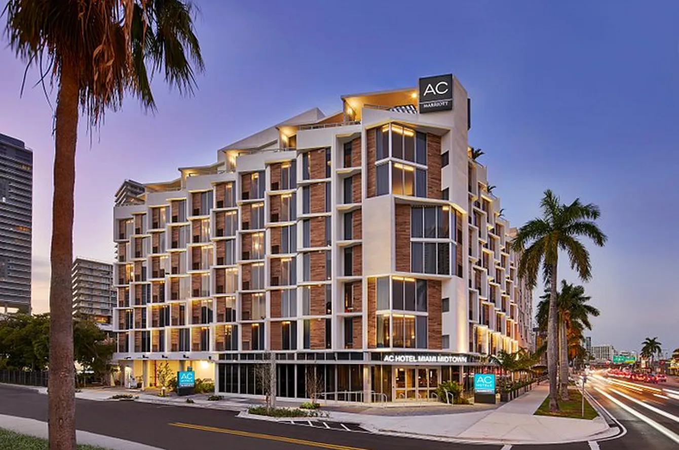 Modern mid-rise AC Hotel Miami Midtown building at dusk with palm trees and street traffic.