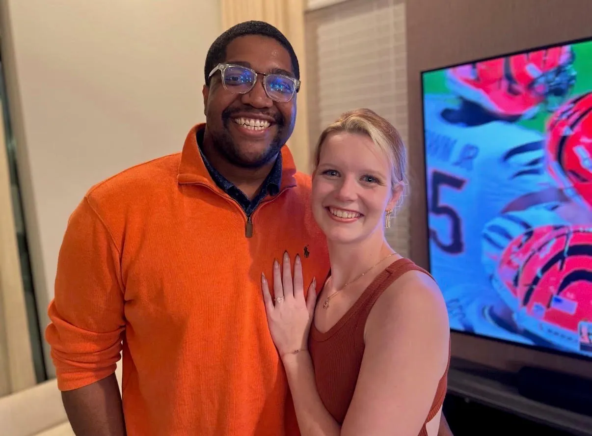 Two smiling people posing together indoors near a colorful TV display.