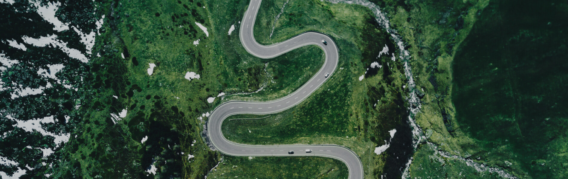 Luftaufnahme einer kurvenreichen Straße mit Fahrzeugen durch grüne bergige Landschaft mit Schneeflecken.