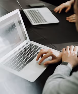 Person typing on a laptop keyboard at a table with another person using a laptop in the background.