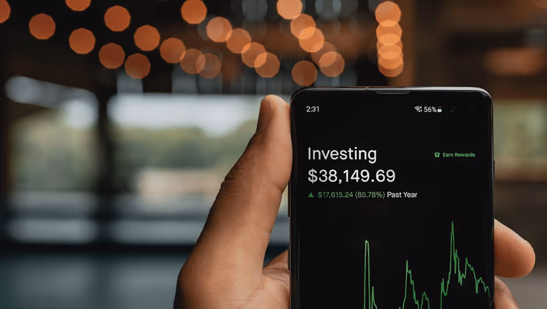 Hand holding phone showing investing app with balance $38,149.69 and 85.78% gain in the past year.