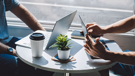 Two people at a round table with laptops and coffee, one person using a tablet device.