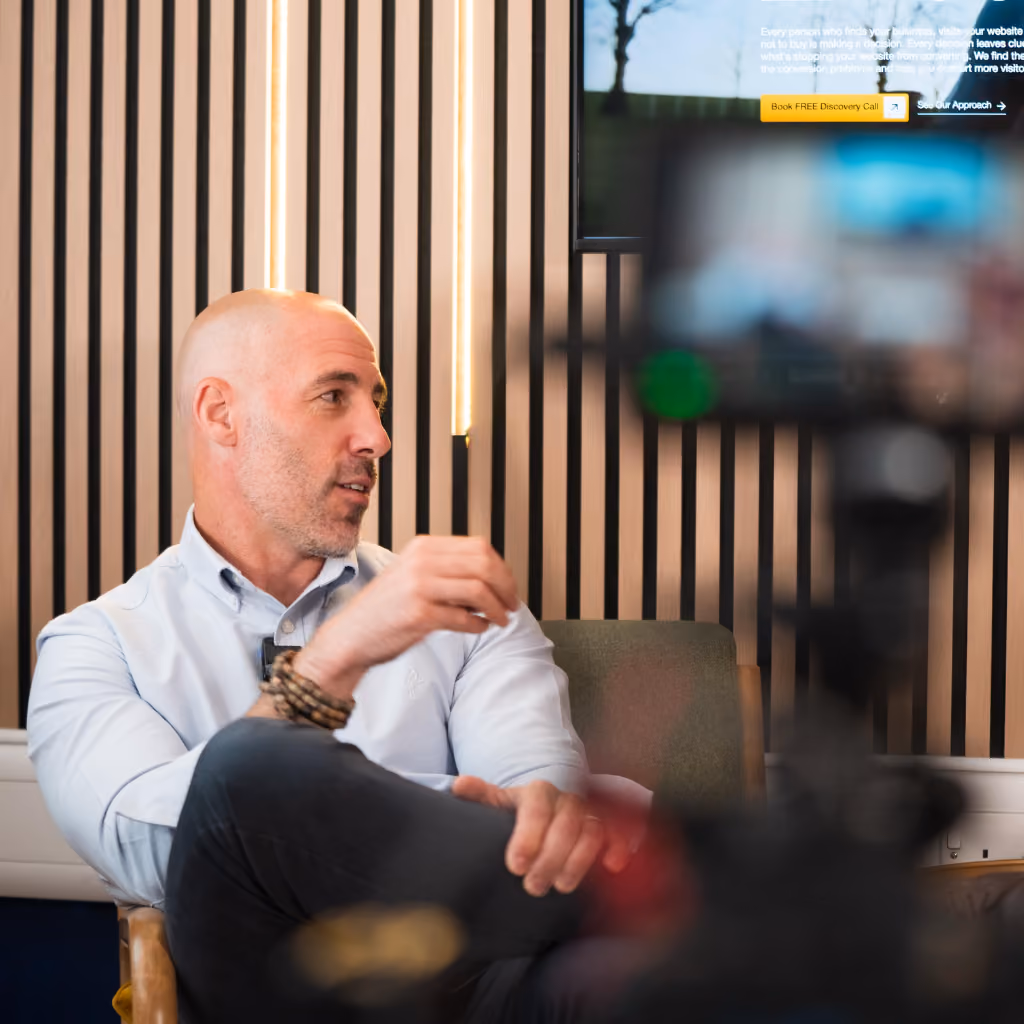 Bald man in white shirt sitting and talking in a modern room with vertical wood panels.