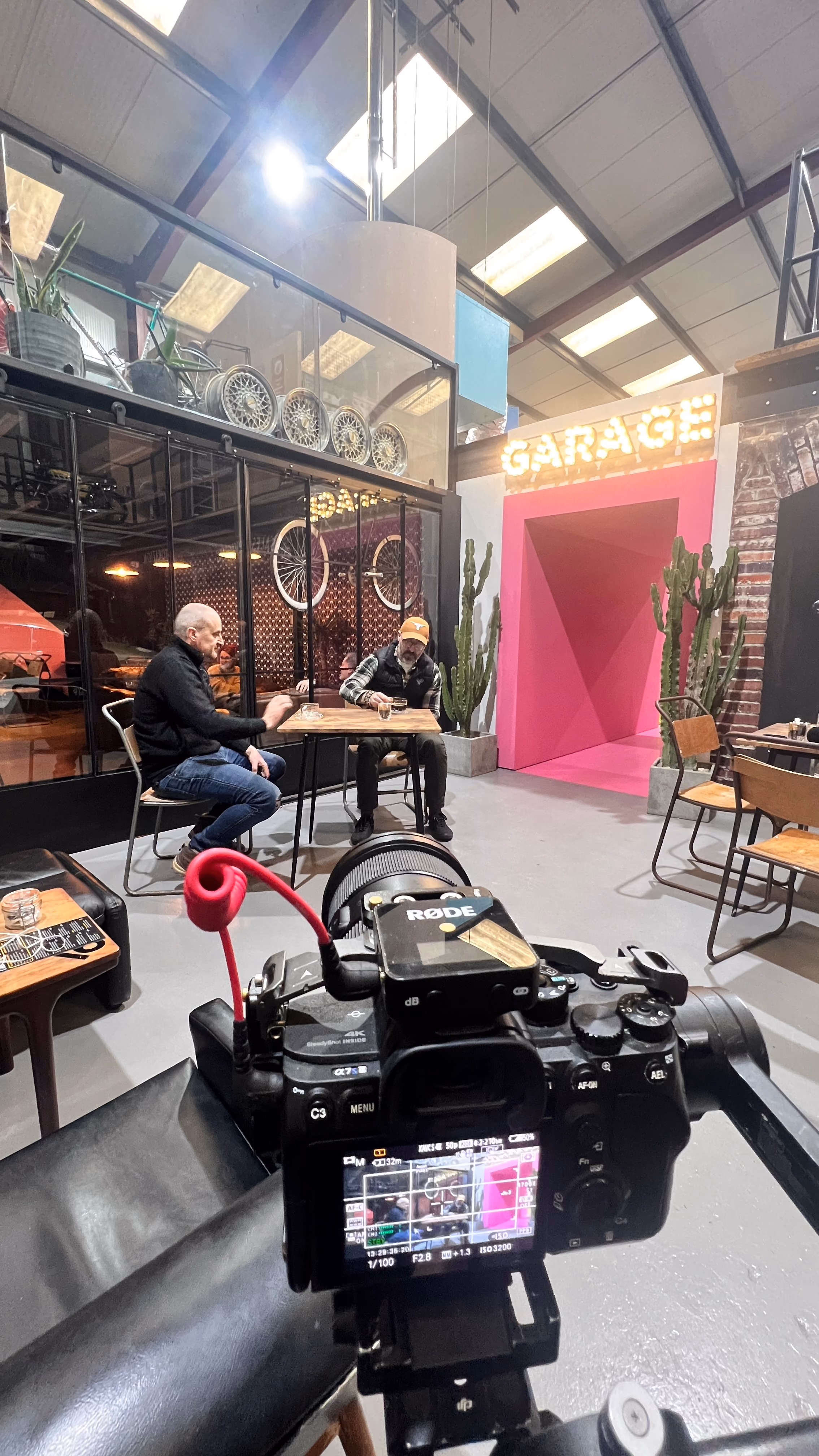 Camera with a RØDE mic filming two men sitting at a table in a garage-themed room with cacti.