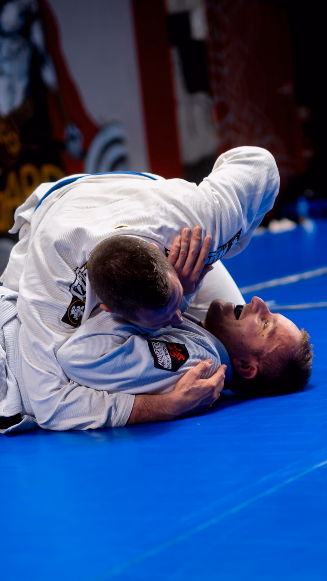 Two men in white martial arts gi grappling on a blue mat during a Brazilian jiu-jitsu match.