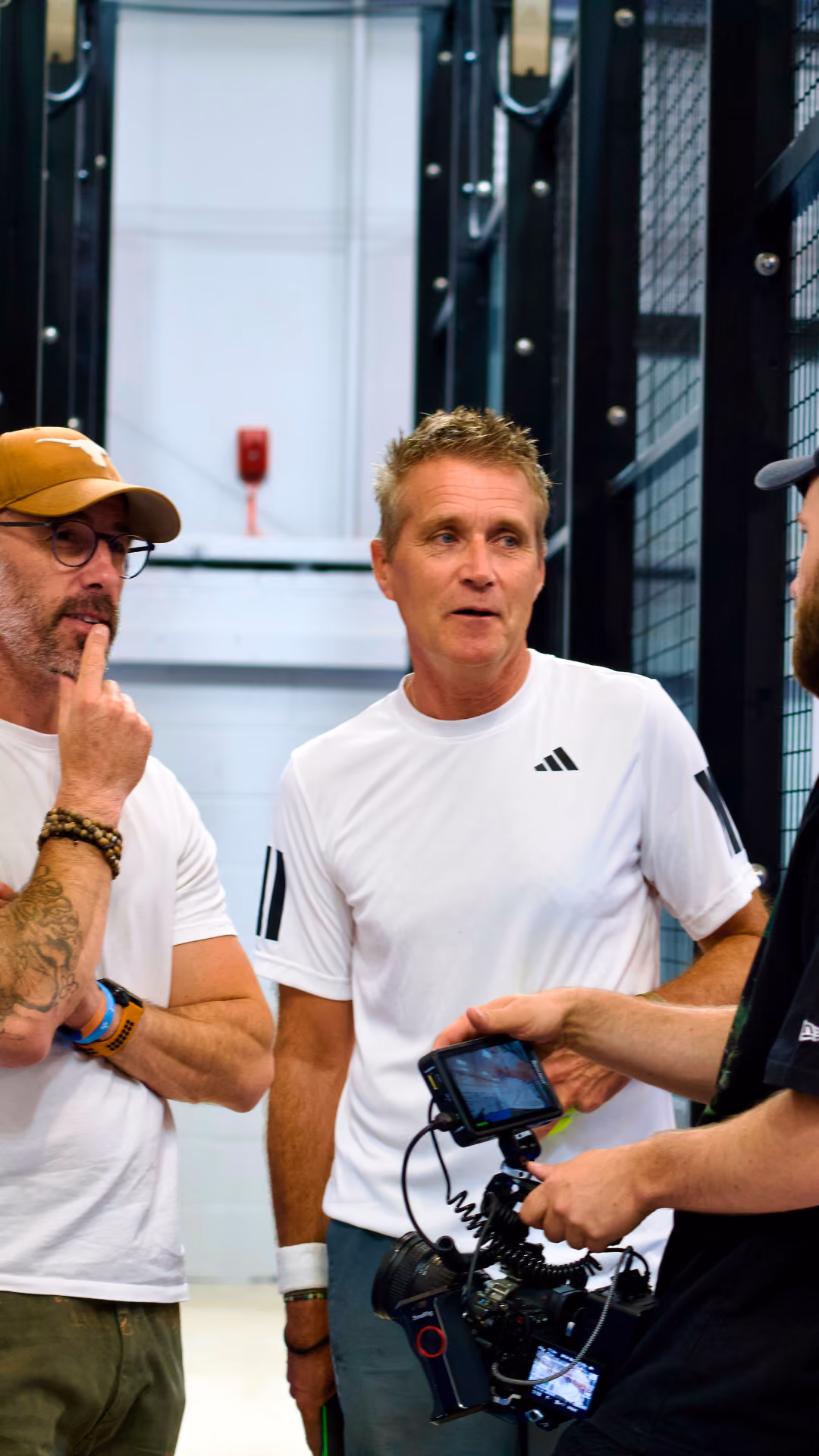 Three men in casual clothes discuss while holding a professional video camera indoors.