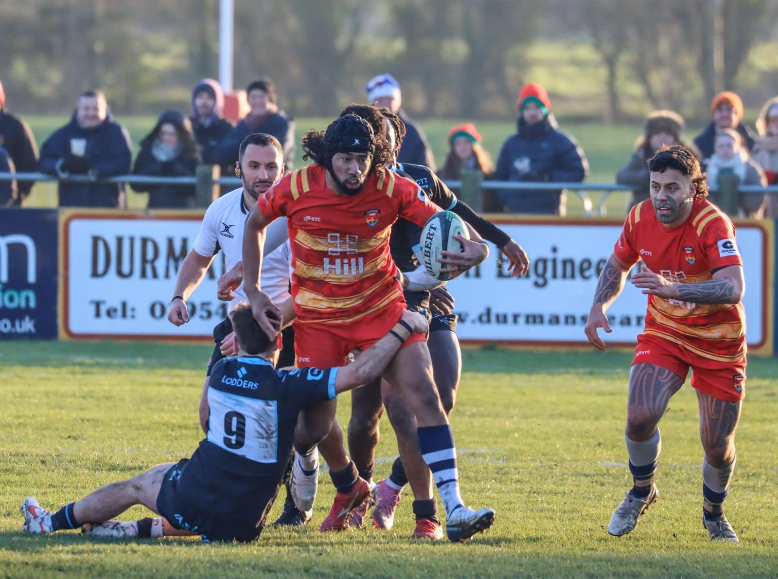 Rugby player in red runs with ball while player in black tries to tackle him on grassy field.