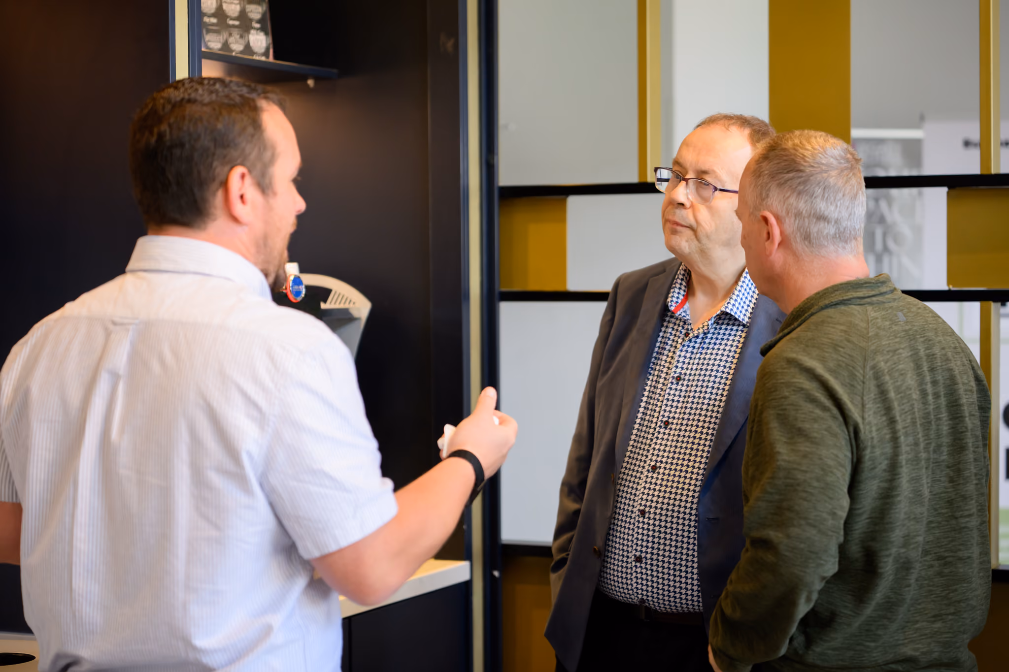 Three men conversing indoors, one in a white shirt speaking, the others listening attentively.
