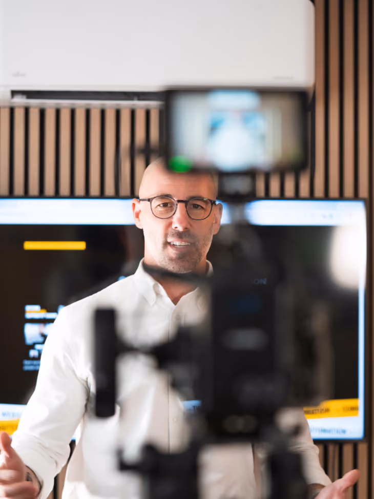 Man in glasses wearing white shirt recording video with a camera in an office setting.