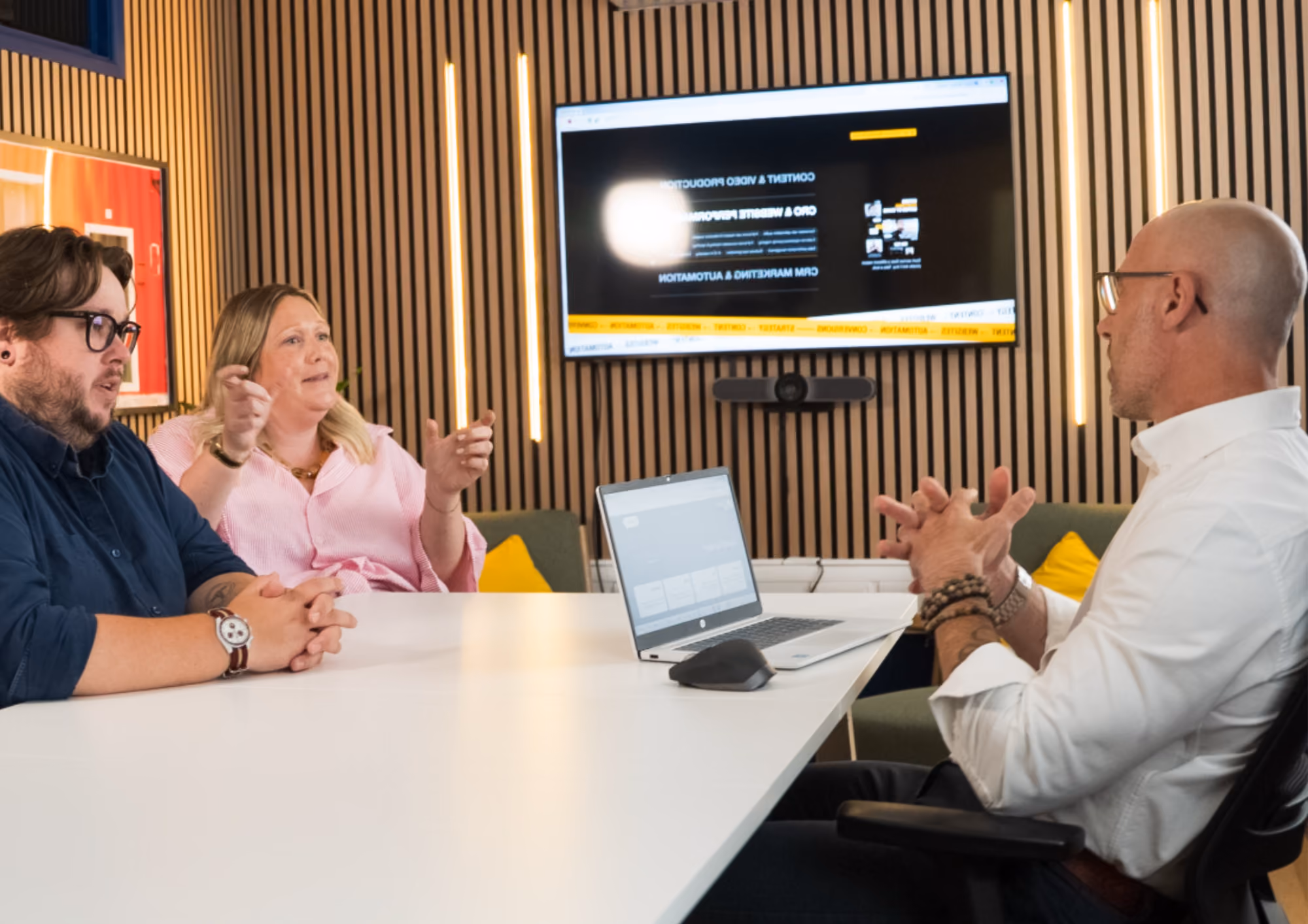 Three colleagues in a meeting room discussing with a laptop and screen on the wall behind them.