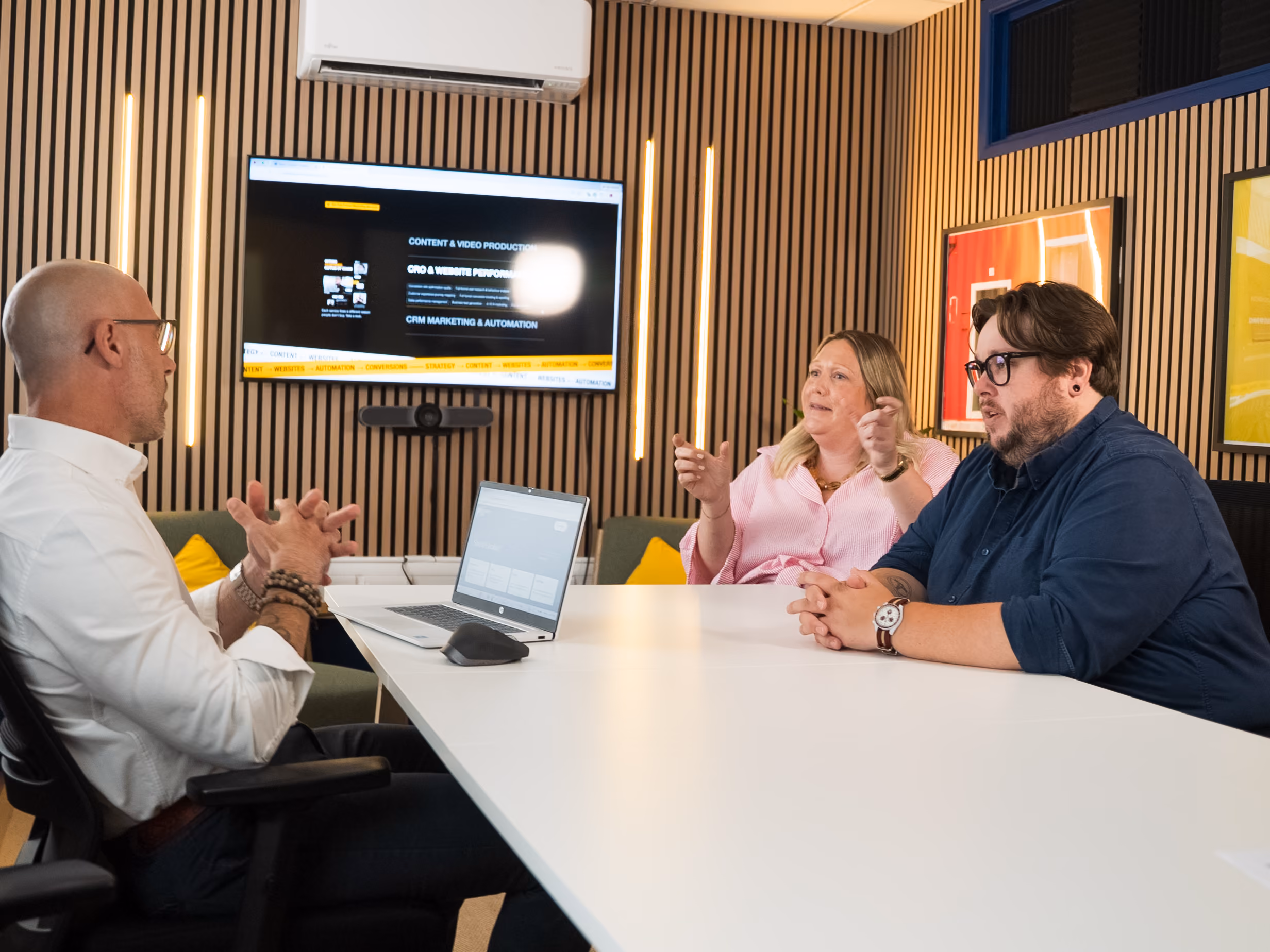 Three colleagues having a meeting at a white table with a screen showing work content on the wall.