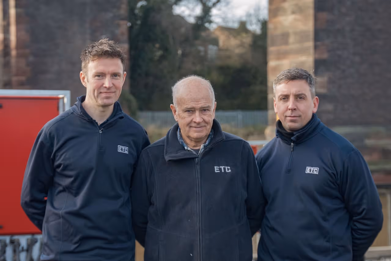 Three men wearing matching navy jackets with ETC logo standing outdoors in front of a blurred industrial background.
