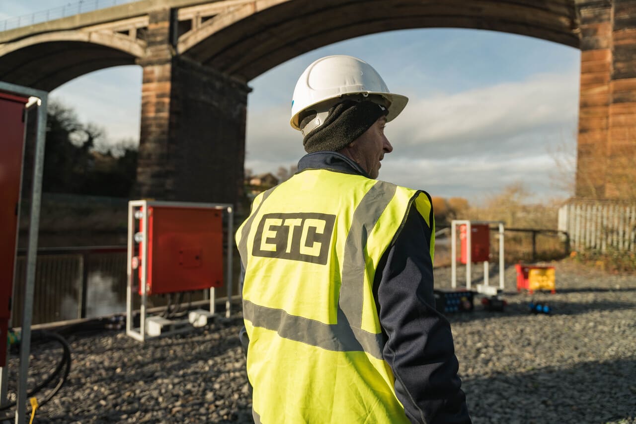 Construction worker wearing a white hard hat and high-visibility vest with ETC logo, standing under a large stone bridge.
