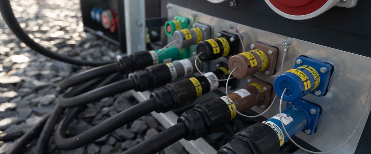 Close-up of multiple heavy-duty industrial cables connected to a metal panel with color-coded plugs and yellow warning labels.