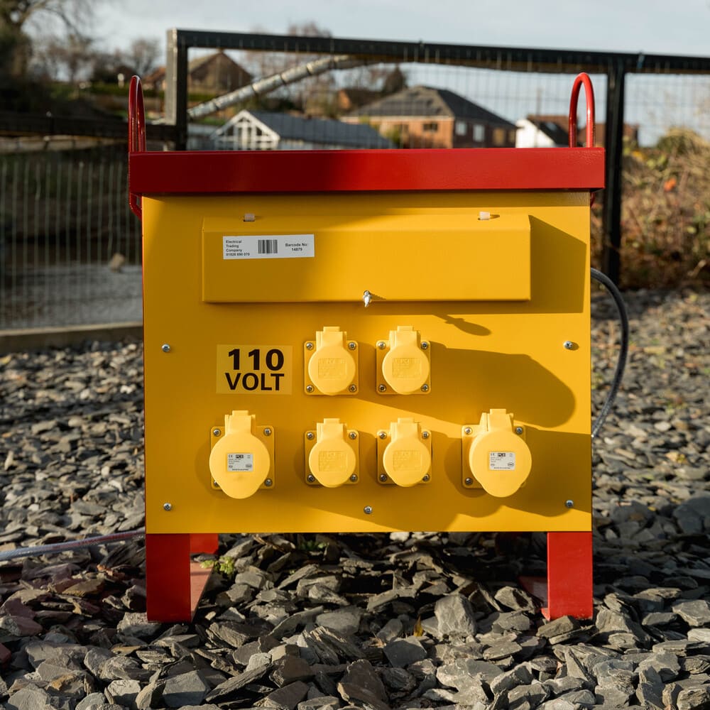 Yellow and red portable electrical power distribution box with six yellow sockets labeled 110 volt on rocky ground outdoors.