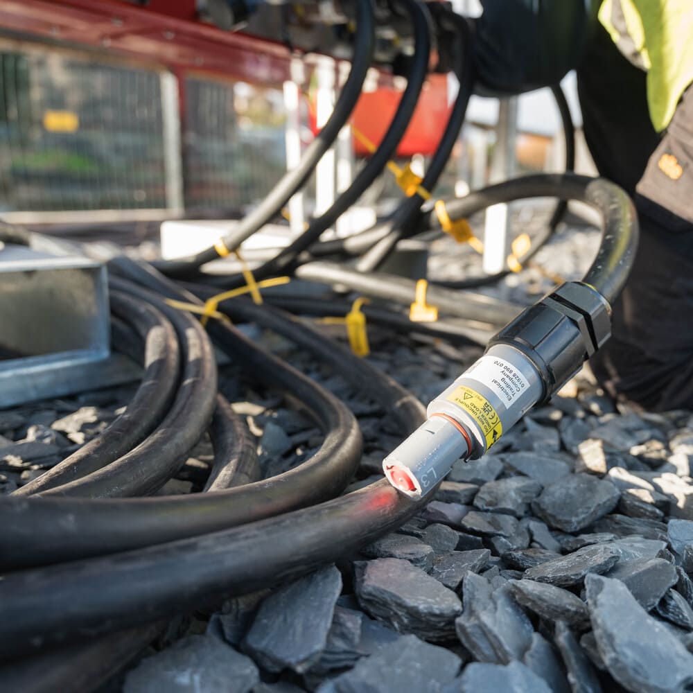 Thick black electrical cables laid on gray gravel with a close-up of a connector labeled with warning and technical information.