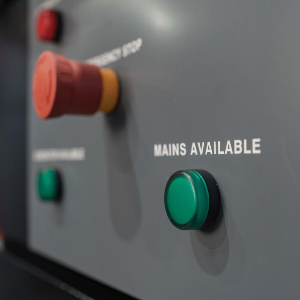 Close-up of a control panel with a green button labeled 'MAINS AVAILABLE' and a red emergency stop button in the background.