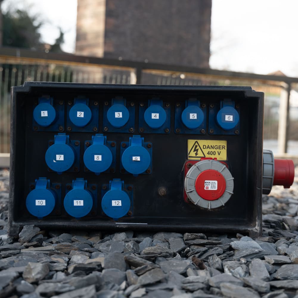 Black electrical panel box with twelve blue numbered sockets and a red circular main output switch, placed on gravel outdoors.