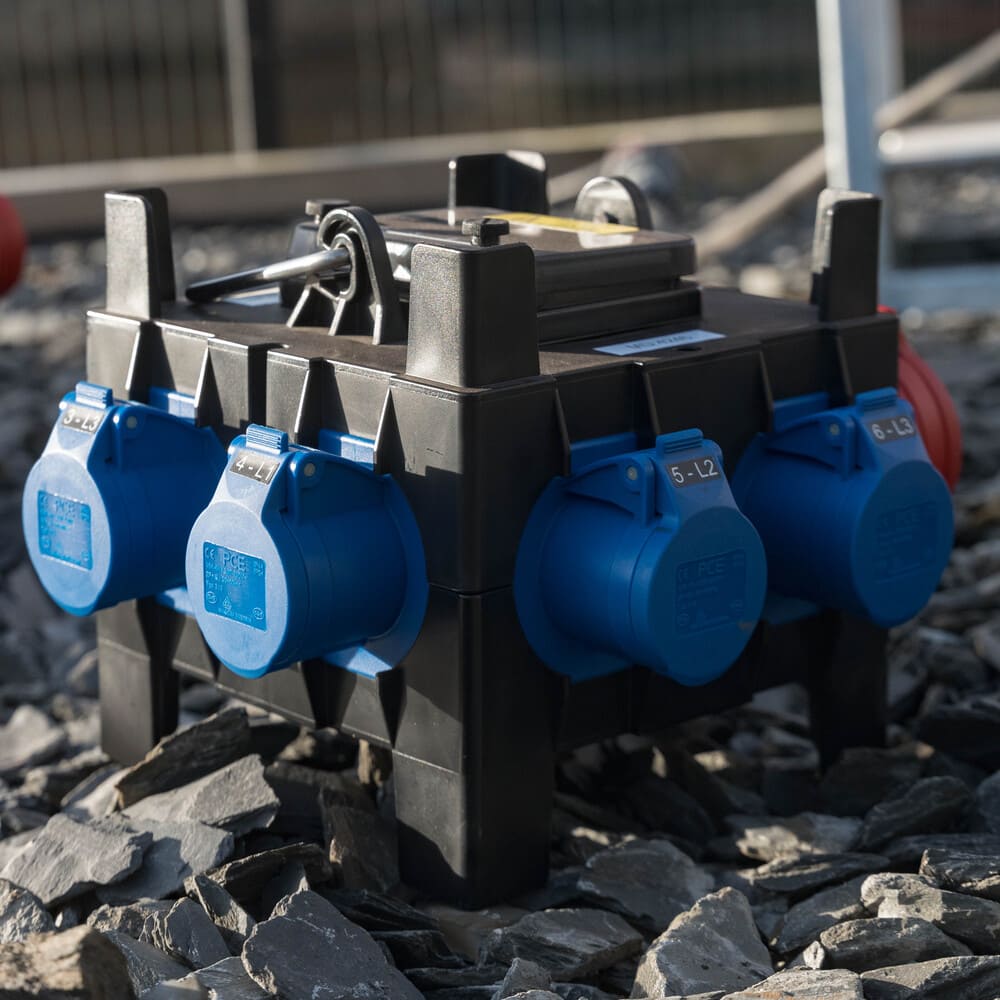 Black power distribution box with six blue industrial sockets placed on rocky ground outdoors.
