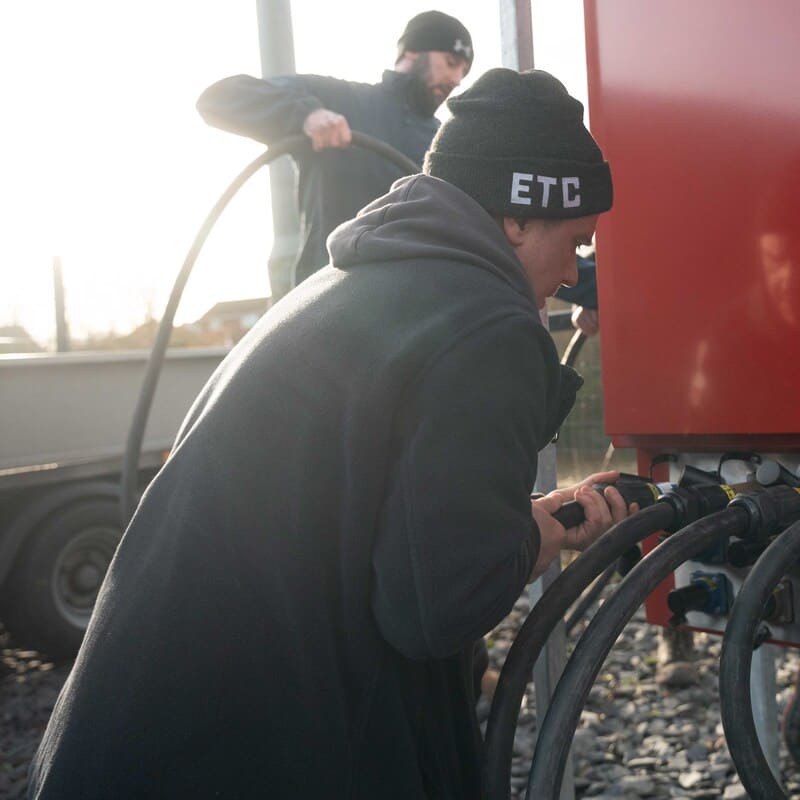 Two men wearing black beanies and jackets are handling thick black hoses connected to a red machine outside on a gravel surface.