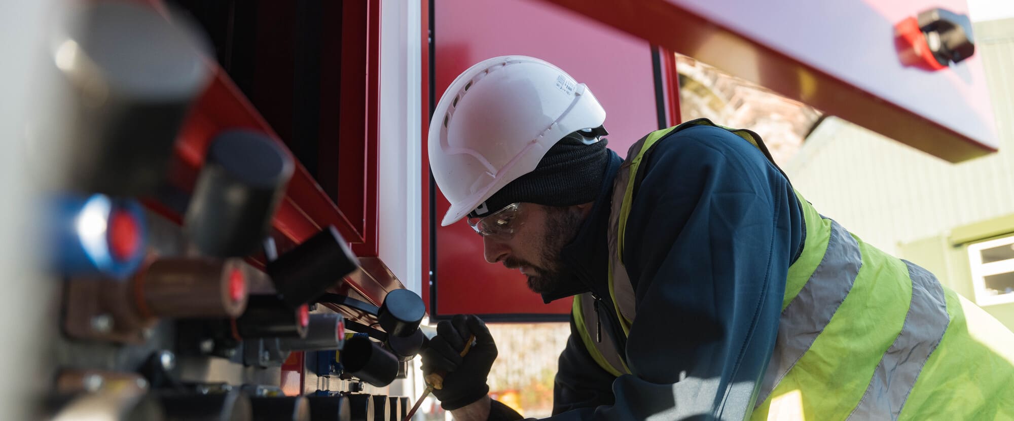 Worker in safety helmet and vest inspecting gauges on a red industrial panel outdoors.
