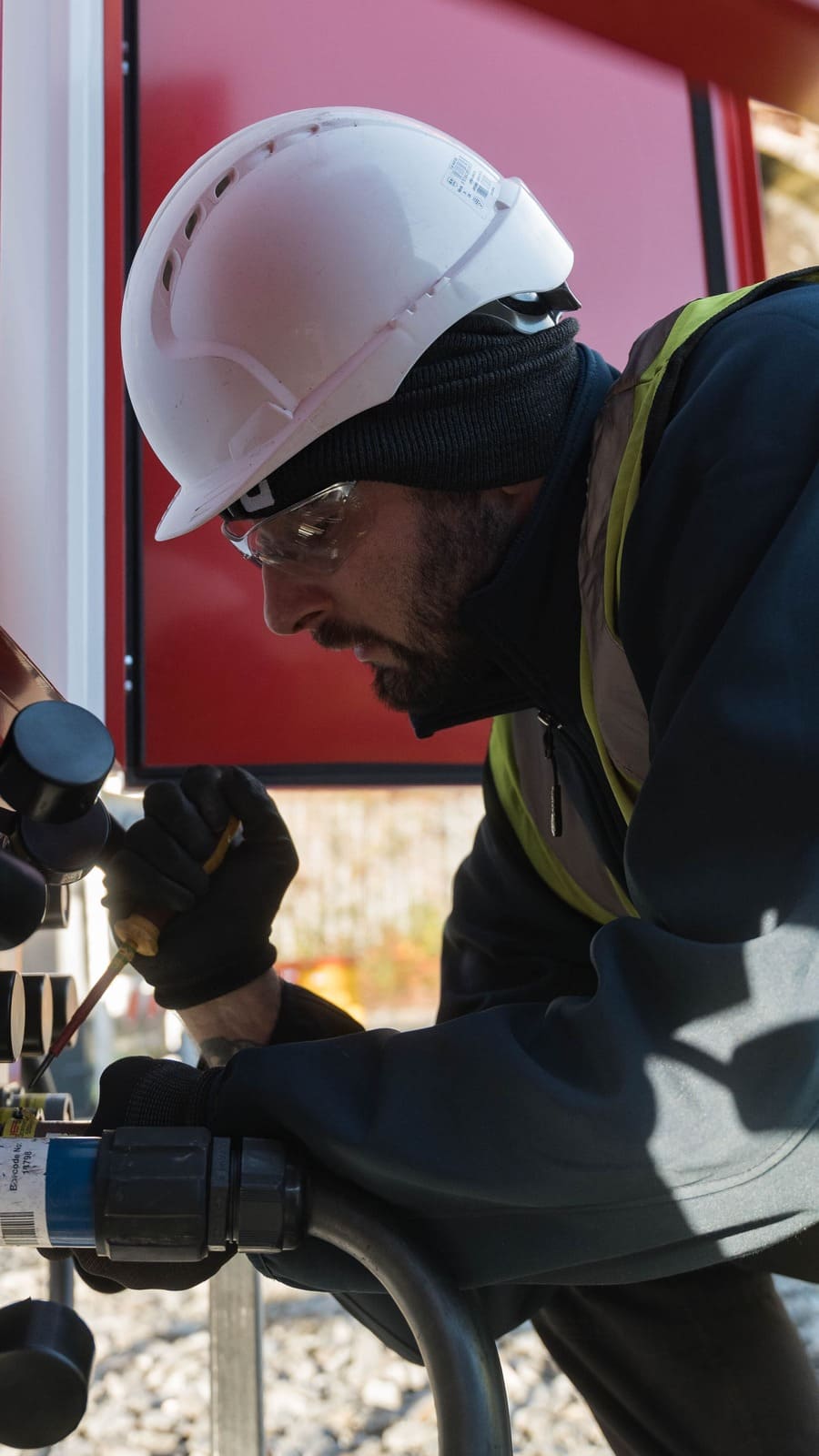 Construction worker wearing a white hard hat and safety glasses, inspecting equipment with a tool.