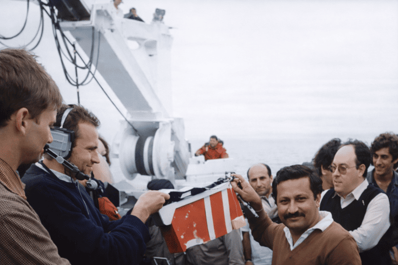 Group of people aboard a boat, with one man wearing a headset holding a red and white striped clapperboard.