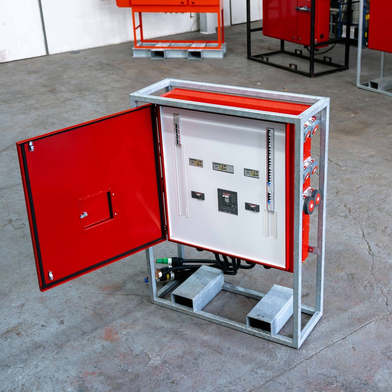 Open industrial electrical control panel with red door and various gauges and controls mounted on a metal frame in a workshop.