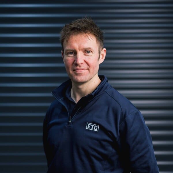 Smiling man wearing a dark blue ETC-branded jacket standing in front of a dark, horizontally ridged background.