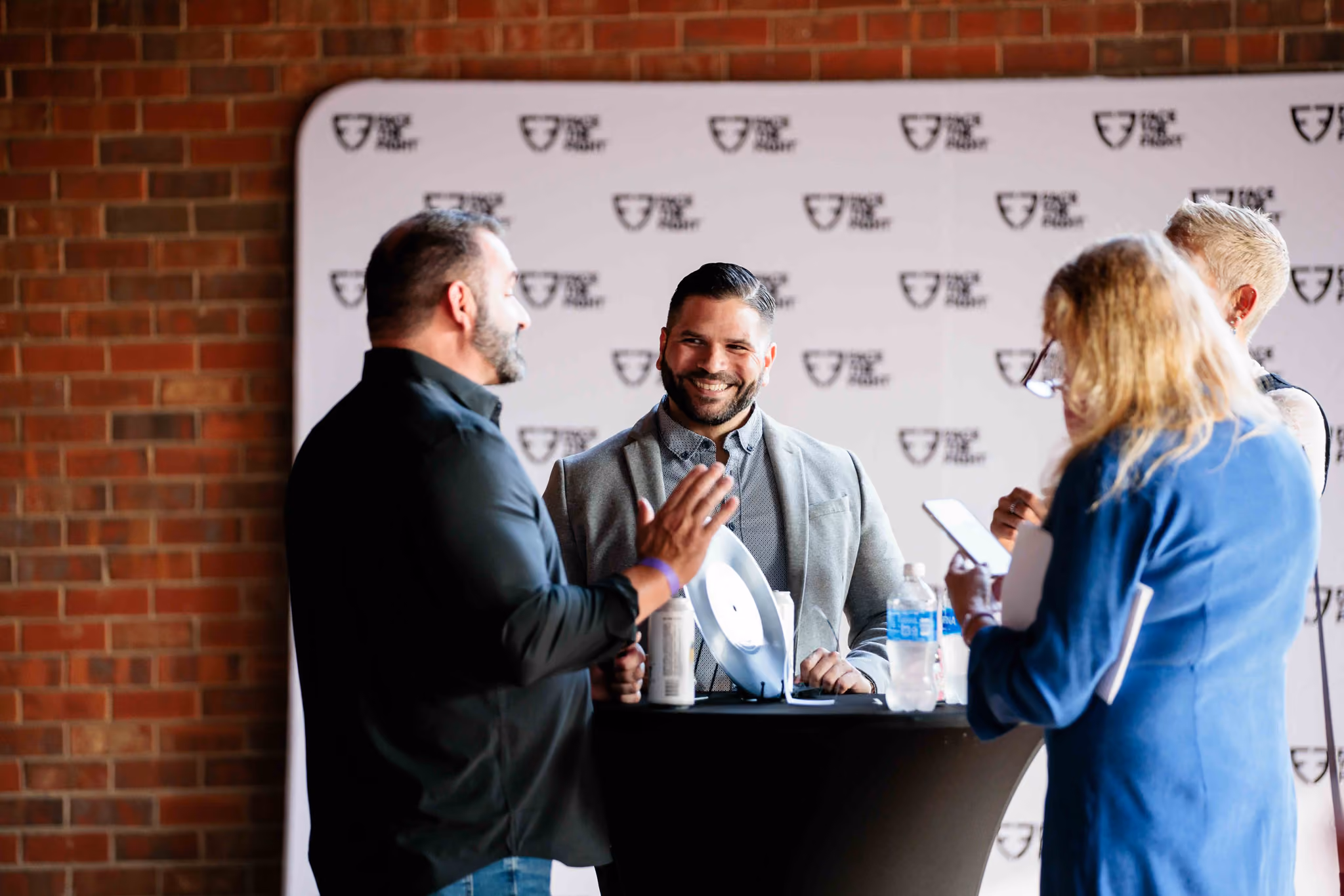 Four people standing around a tall black table engaged in conversation with a brick wall and backdrop behind them at the Face the Fight Grand Ole Opry reception.