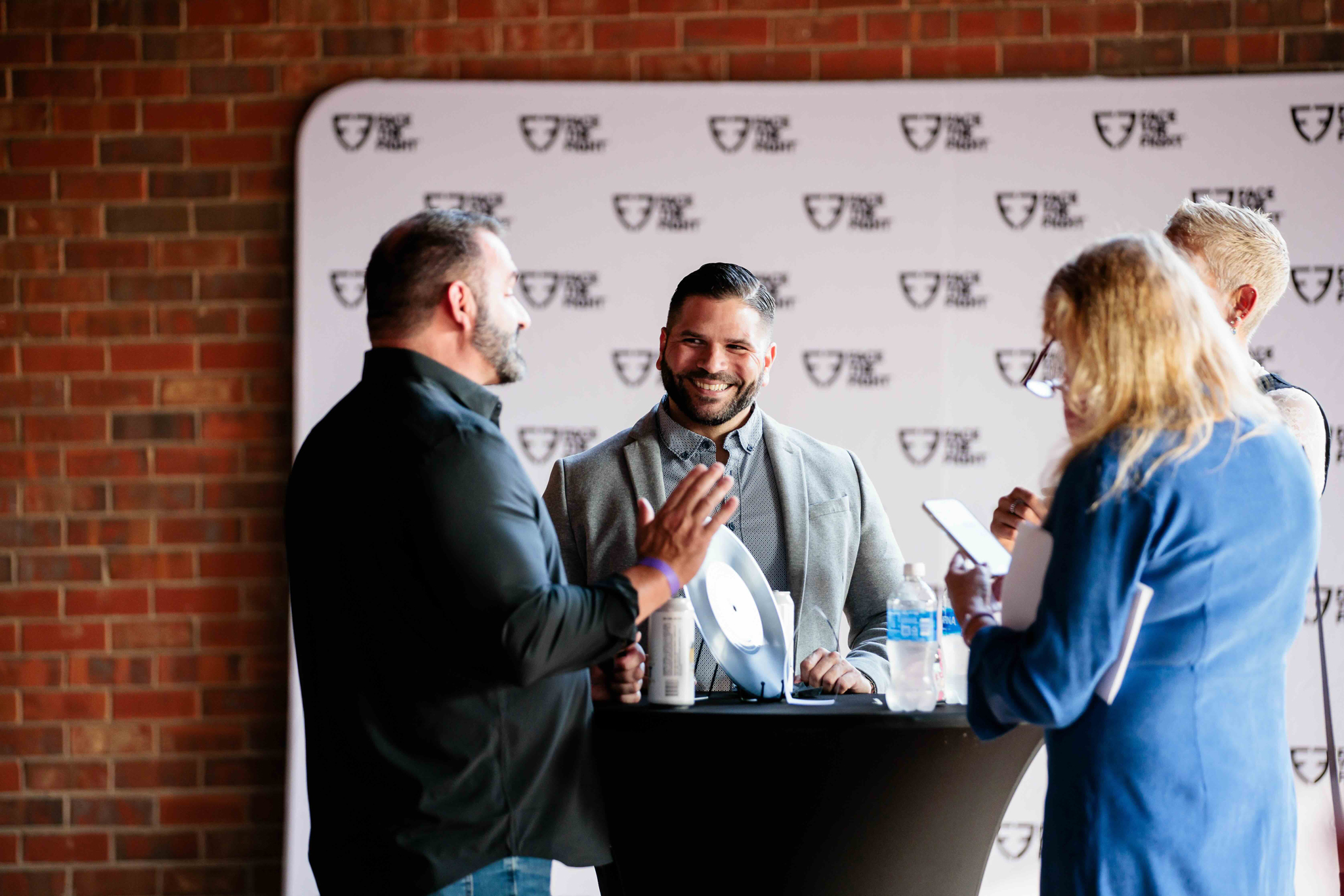 Four people standing around a tall black table engaged in conversation with a brick wall and backdrop behind them at the Face the Fight Grand Ole Opry reception.