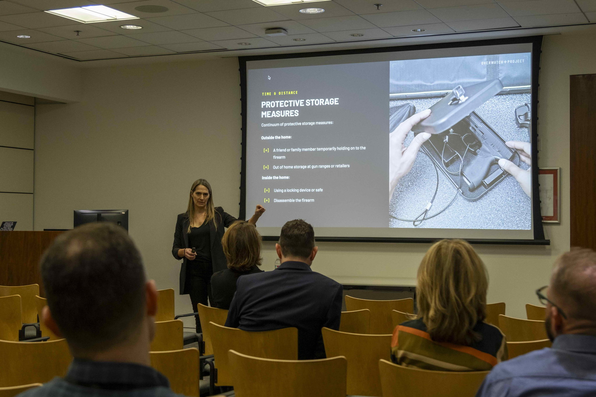 A woman giving a presentation on protective storage measures for firearms in a lecture room with four seated attendees.