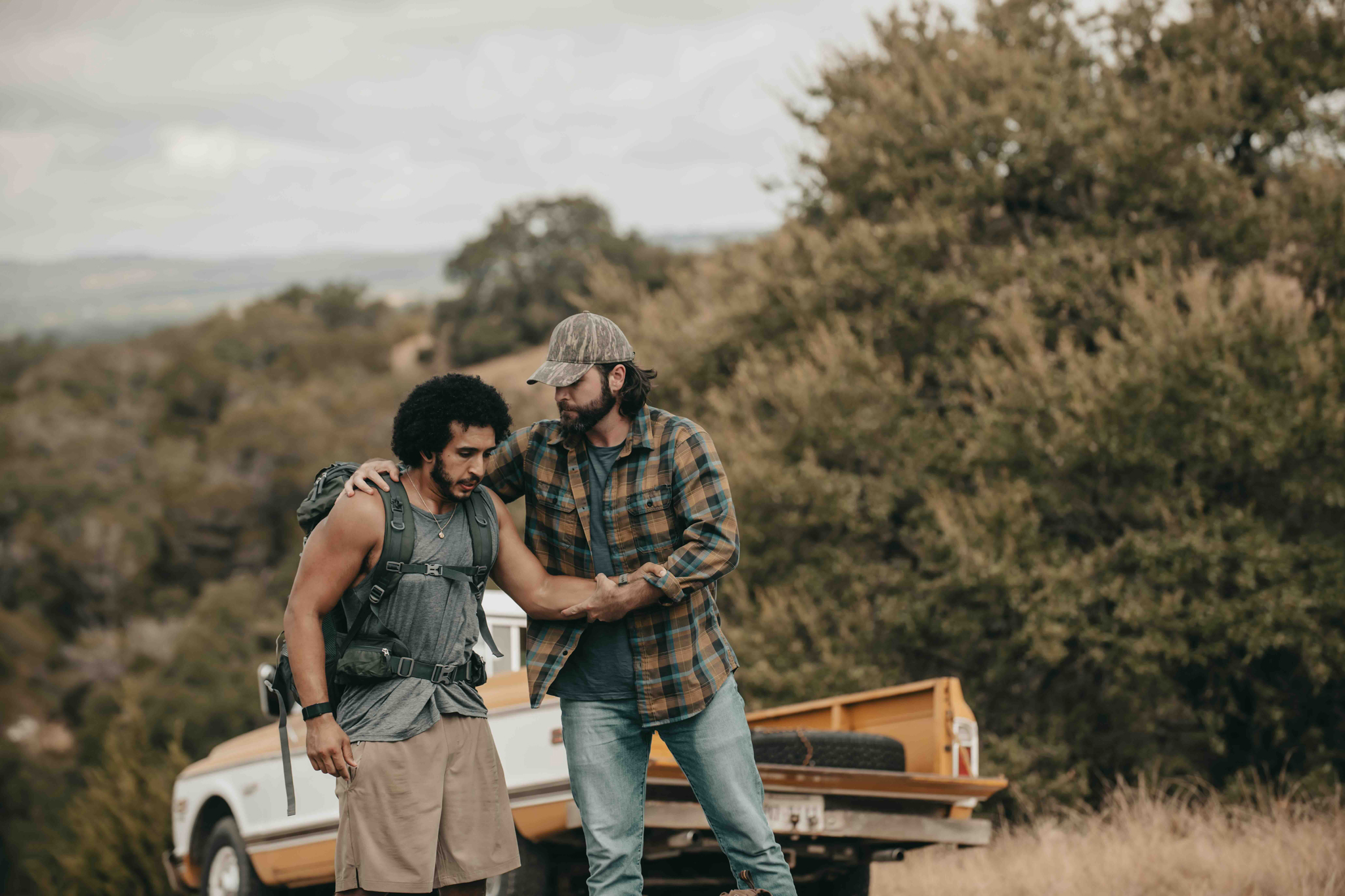 Man with backpack being assisted by another man wearing a plaid shirt and cap near a truck in a wooded area.