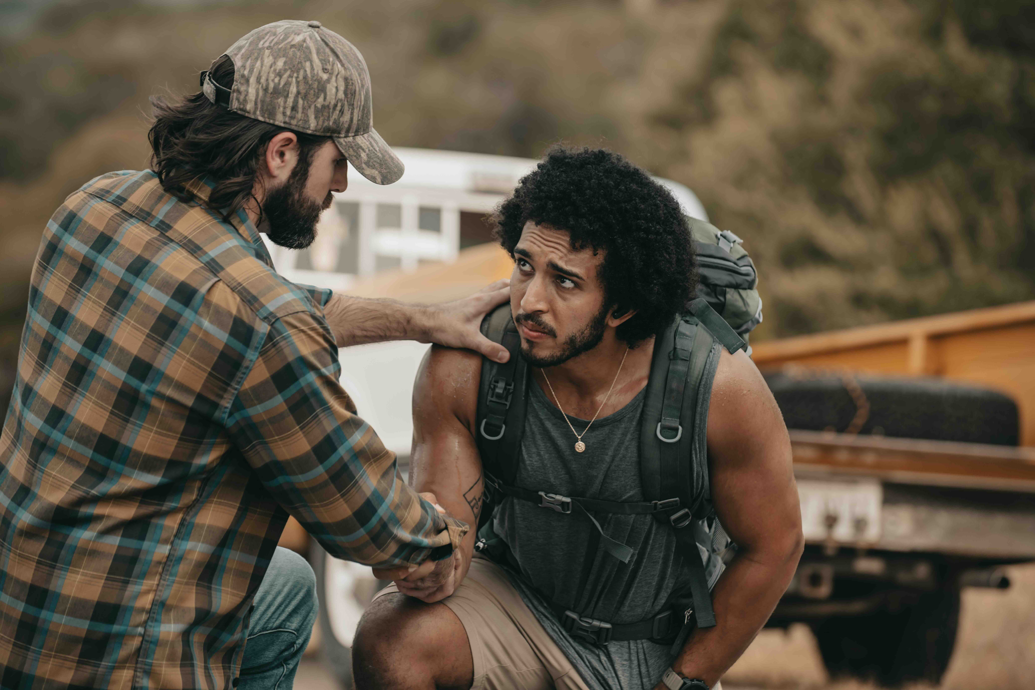 Man wearing a plaid shirt and camo cap helps a tired hiker with backpack and tank top outdoors.