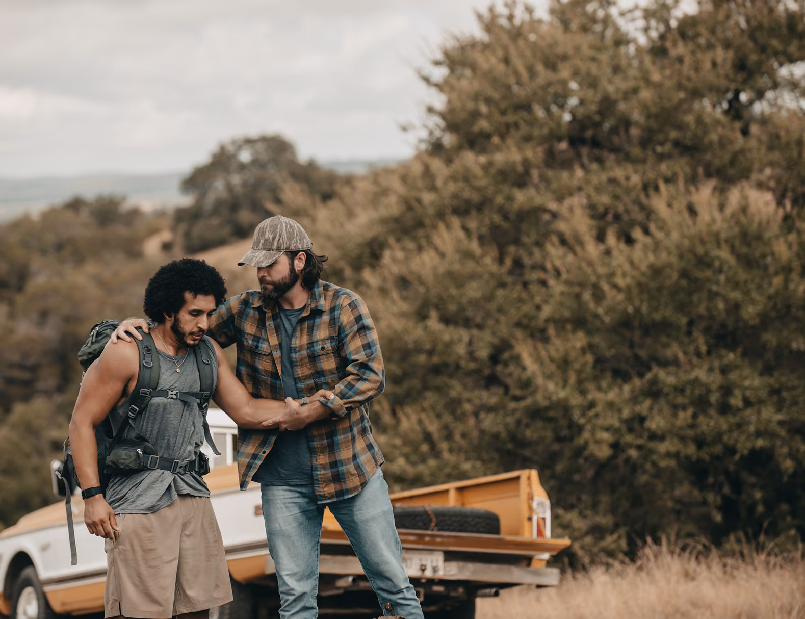 Man in plaid shirt helps another man wearing hiking gear outdoors near a pickup truck.