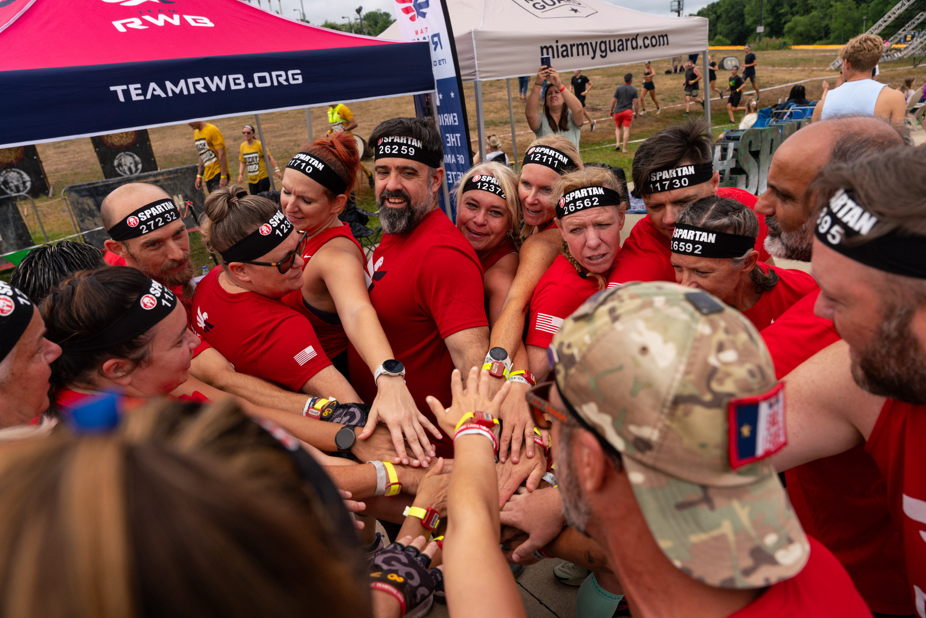 Group of people from Team Re, White, and Blue wearing red shirts and black Spartan headbands putting their hands together in a team huddle at an outdoor event.