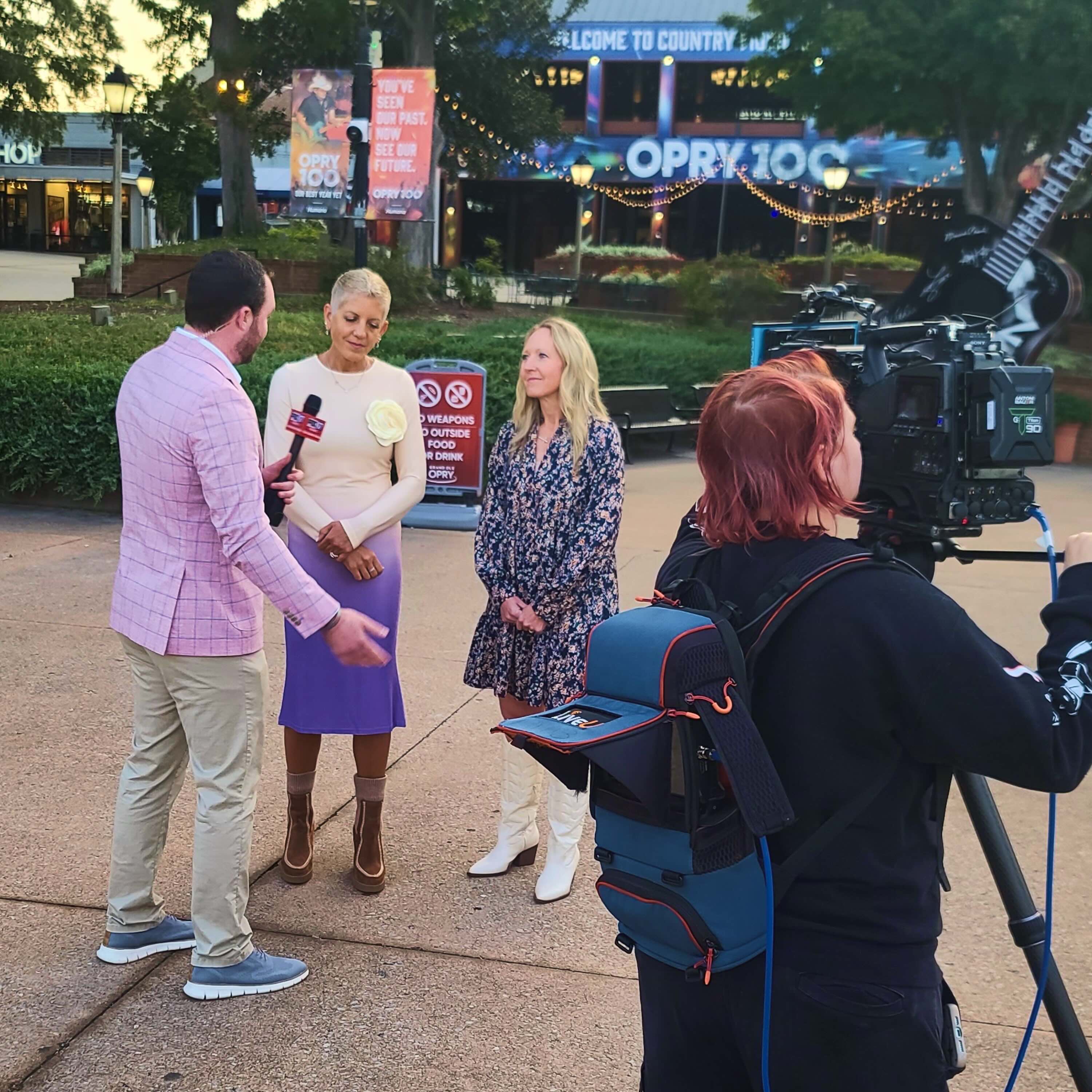 Outdoor interview with a man holding a microphone speaking to two women, while a camerawoman films the scene near Opry 100 venue.