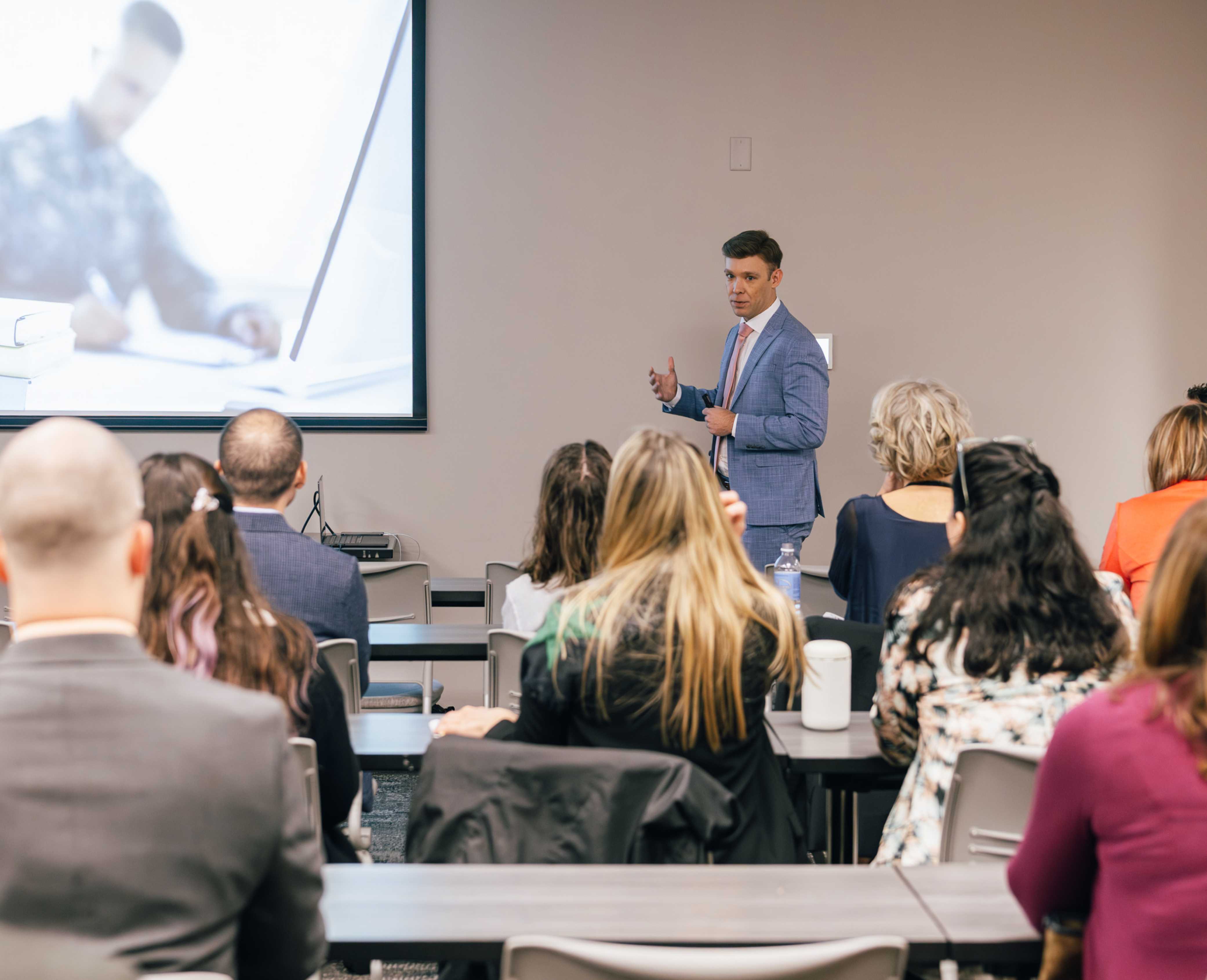 Man in a blue suit giving a presentation to an audience seated at tables, with a projected image on a screen.