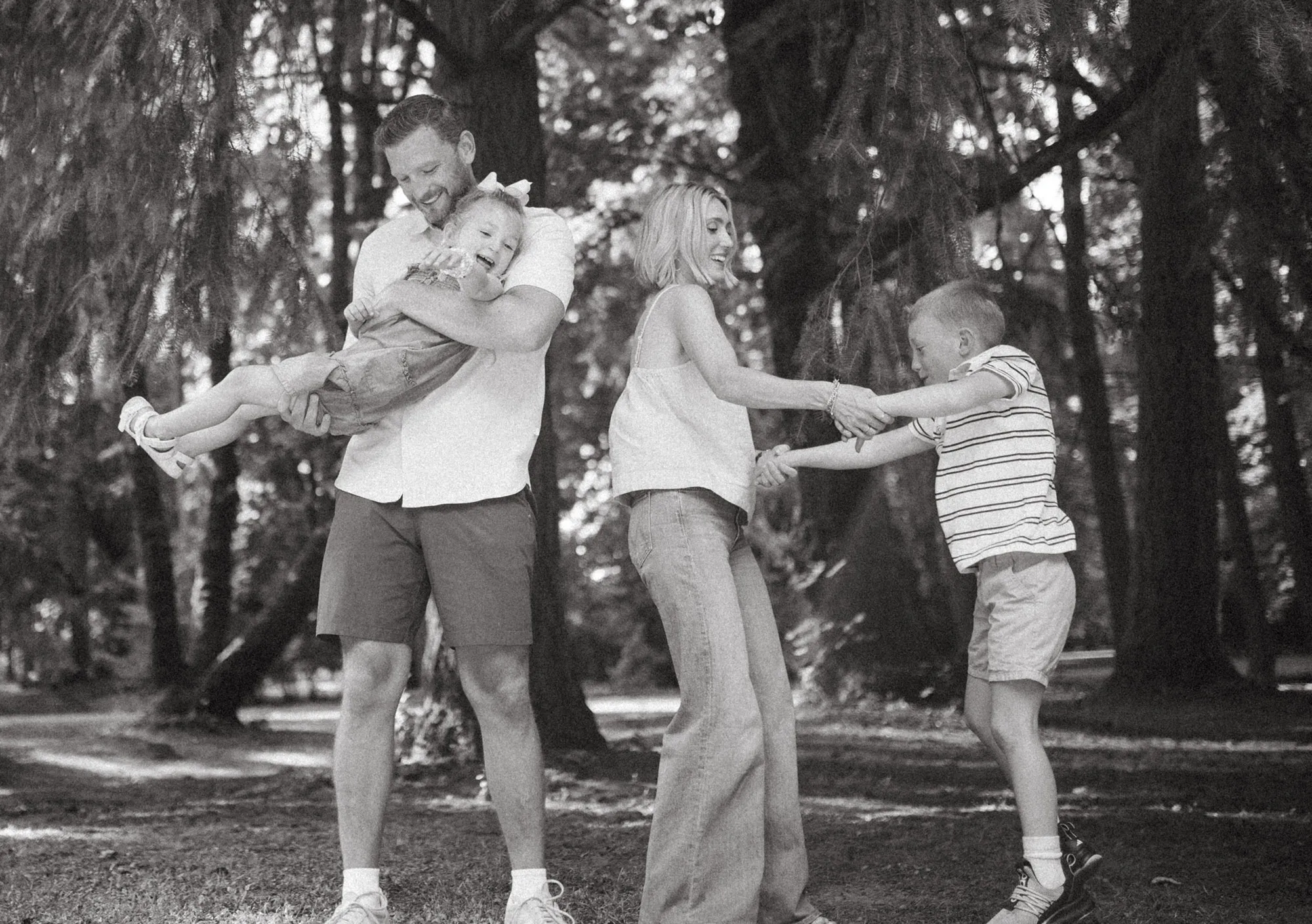 Black-and-white photo of a father holding a smiling daughter while the mother holds hands and plays with their son in a forested park.