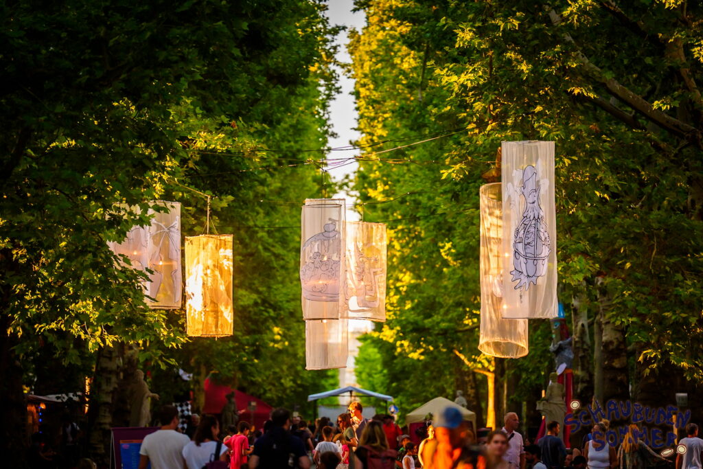 Menschen gehen zum Schaubudensommer durch die schön dekorierte Hauptstraße im Abendlicht.