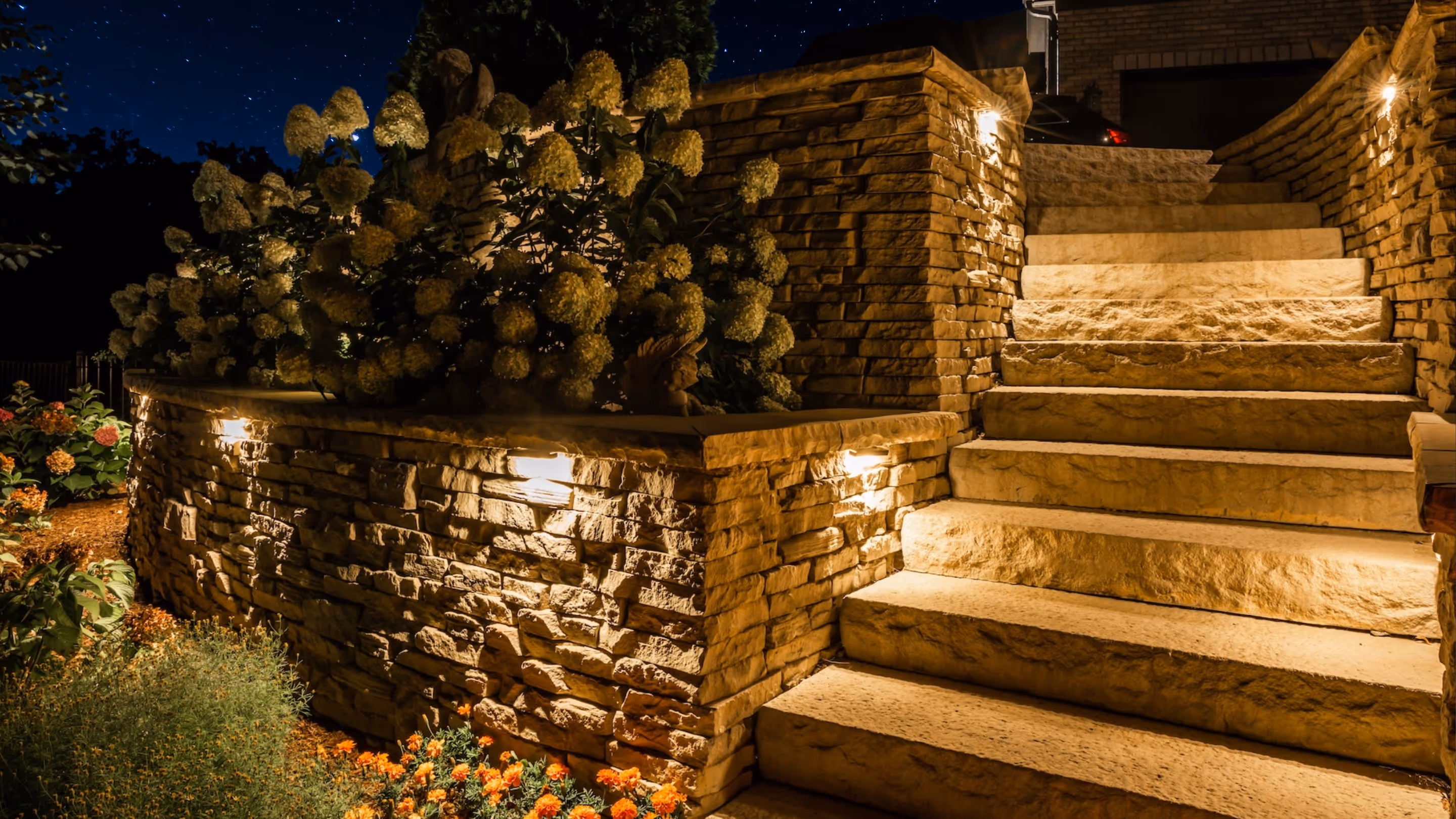 Stone outdoor steps and retaining wall illuminated by warm lights at night with flowering plants nearby.